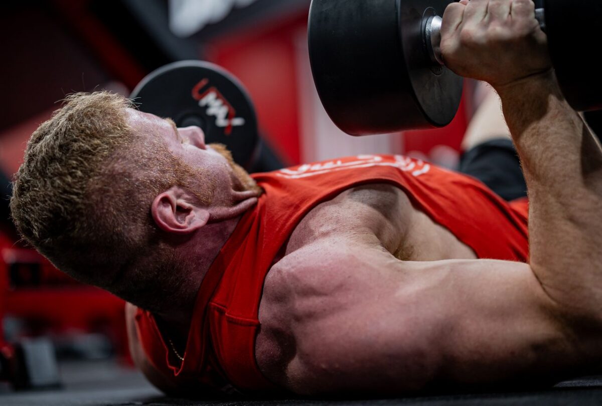 Man lying on a gym floor with dumbbells held above his chest and one upper arm near the floor.