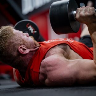Man lying on a gym floor with dumbbells held above his chest and one upper arm near the floor.