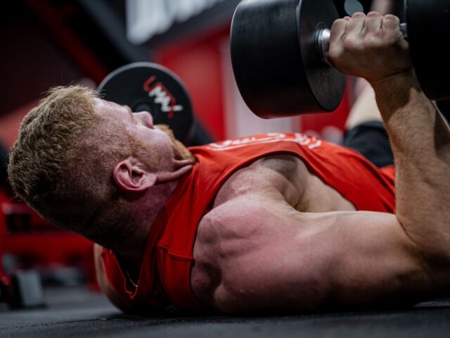 Man lying on a gym floor with dumbbells held above his chest and one upper arm near the floor.