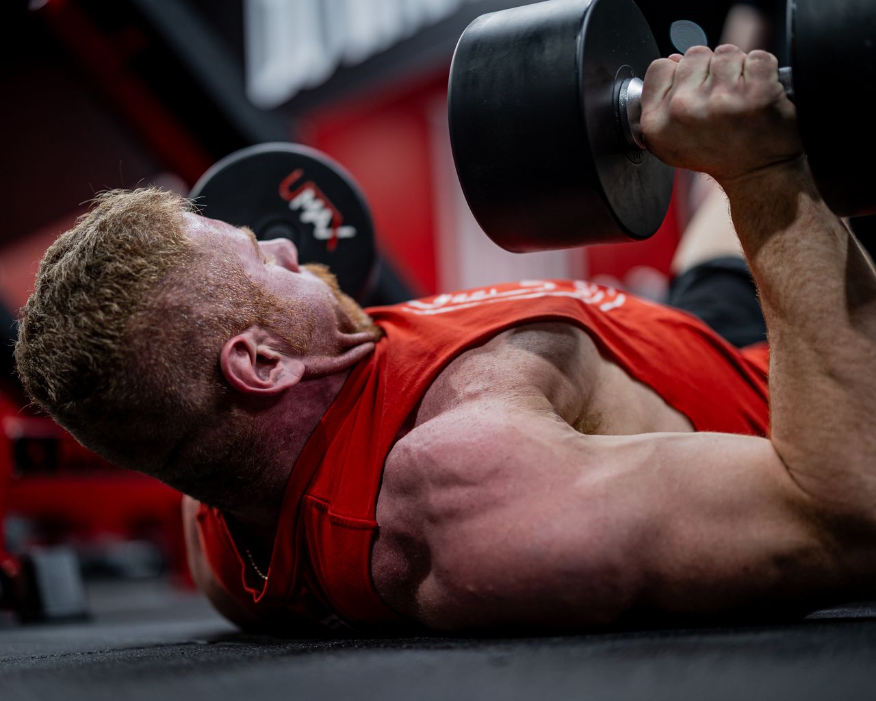 Man lying on a gym floor with dumbbells held above his chest and one upper arm near the floor.