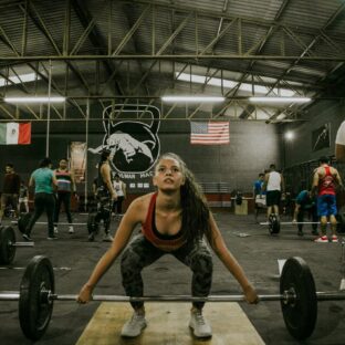 Woman setting up at a loaded barbell while other athletes train in a CrossFit gym.