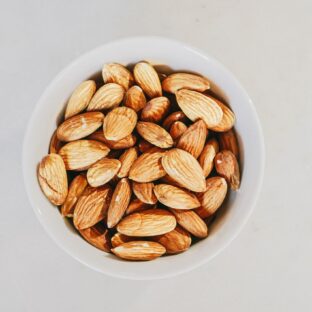 Whole almonds in a white bowl on a pale surface, viewed from above.