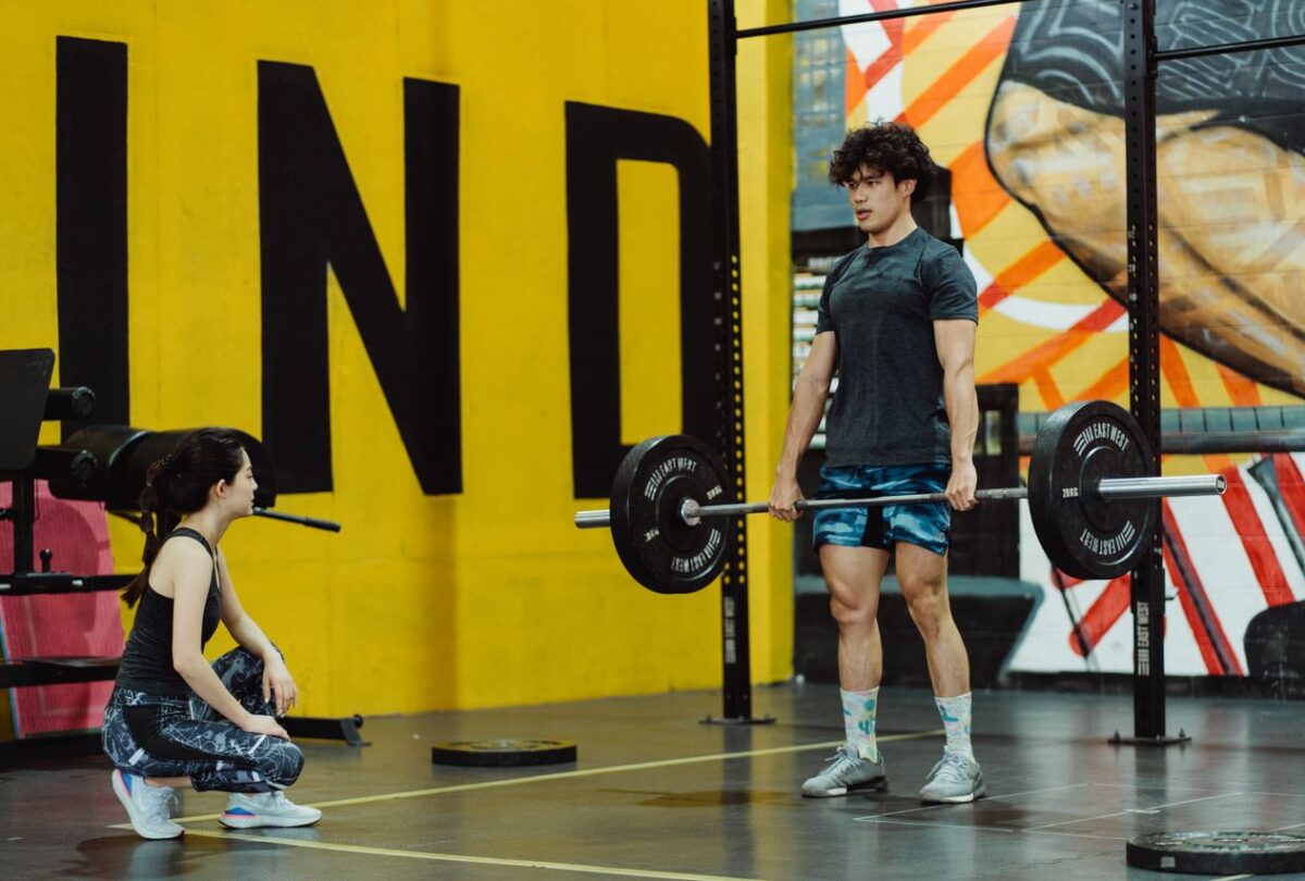 Man standing upright with a barbell held at thigh height while a trainer crouches beside him.