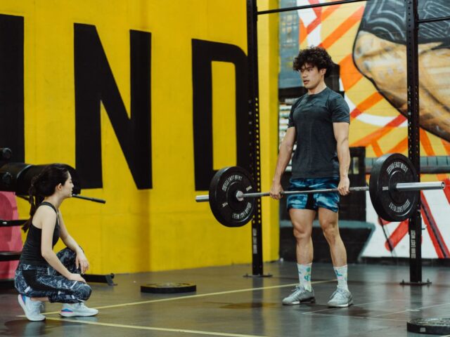 Man standing upright with a barbell held at thigh height while a trainer crouches beside him.