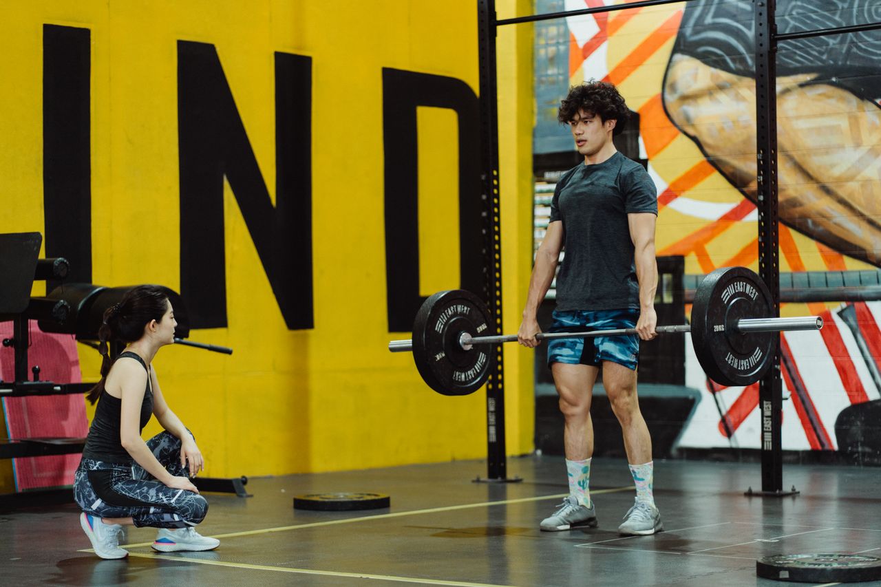 Man standing upright with a barbell held at thigh height while a trainer crouches beside him.