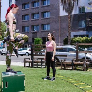 Woman holding dumbbells with one foot planted on a box and the other knee raised, while another woman watches.