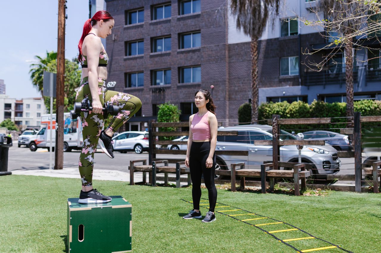 Woman holding dumbbells with one foot planted on a box and the other knee raised, while another woman watches.