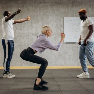 Woman performing an unweighted squat in a gym, with two other people nearby.