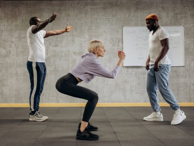 Woman performing an unweighted squat in a gym, with two other people nearby.
