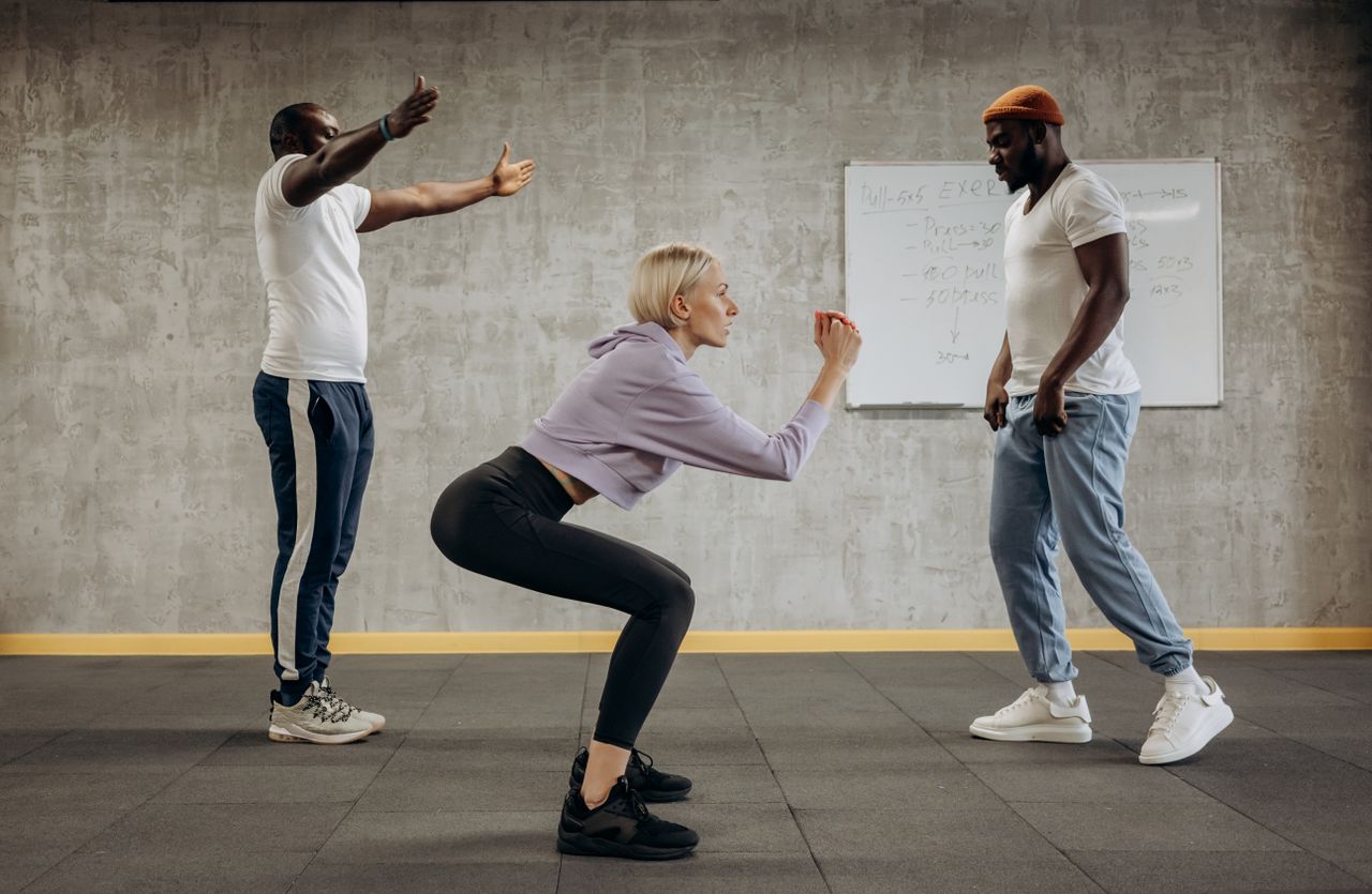 Woman performing an unweighted squat in a gym, with two other people nearby.