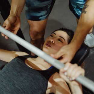 Woman bench pressing a barbell on a flat bench with a spotter assisting above her chest.