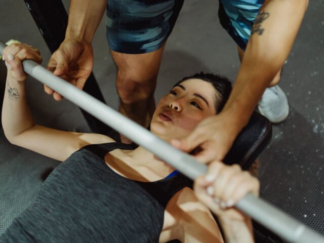Woman bench pressing a barbell on a flat bench with a spotter assisting above her chest.