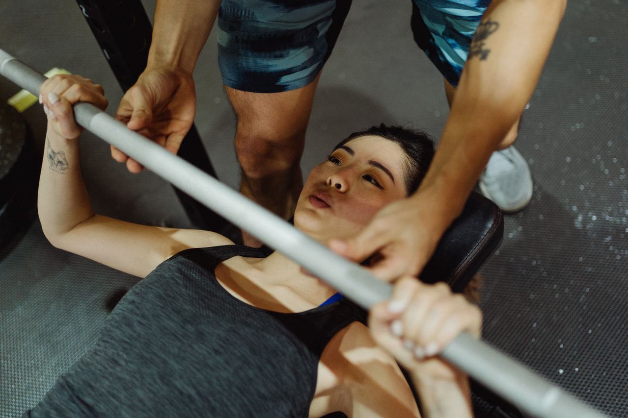 Woman bench pressing a barbell on a flat bench with a spotter assisting above her chest.