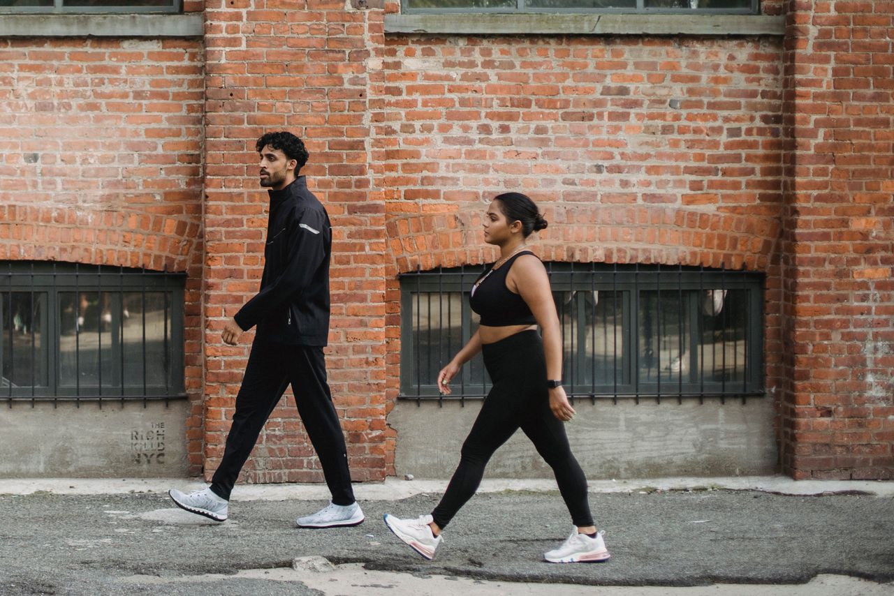 Two adults in athletic clothing walking along a sidewalk beside a brick building.
